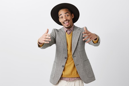 Studio shot of positive friendly african-american man in trendy outfit and hat pulling hands towards camera, inviting mates to come inside, welcoming guest to partyの写真素材