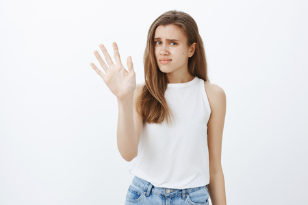 Portrait of displeased unhappy european girl with blond hair in stylish outfit, raising palm in stop or enough gesture, rejecting disturbing offer, expressing indifference and dislikeの写真素材