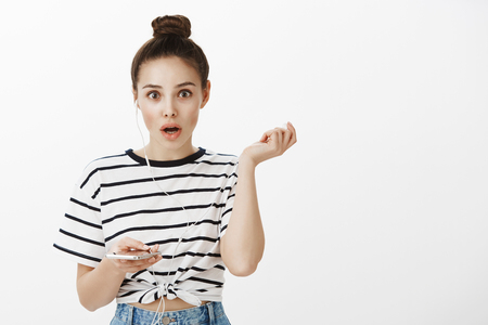 Girl is shocked hearing amazing news, interrupted of listening music in earbuds. Amazed good-looking feminine woman with bun, taking off earphones and holdign smartphone, dropping jaw from surpriseの写真素材