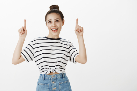 Girl feels great walking under sun in good weather. Portrait of charming attractive european woman with bun hairstyle, being excited and happy while pointing upwards, standing over white wallの写真素材