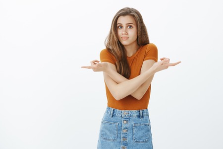 Unsure doubtful attractive Caucasian girl with fair hair, pointing in different directiong with arms crossed on chest, shrugging, being unaware and confused in what direction choose over gray wallの写真素材