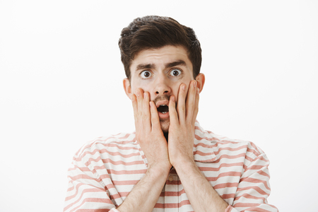 Studio shot of shocked and anxious handsome male model with moustache, touching cheeks with hands nervously, dropping jaw and staring at camera, being stunned with disappointing rumor or newsの写真素材