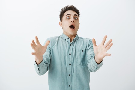 Indoor shot of stunned funny caucasian male with pale face, tilting backwards and pulling raised palms towards camera, shouting, being shocked and afraid, gasping over gray backgroundの写真素材