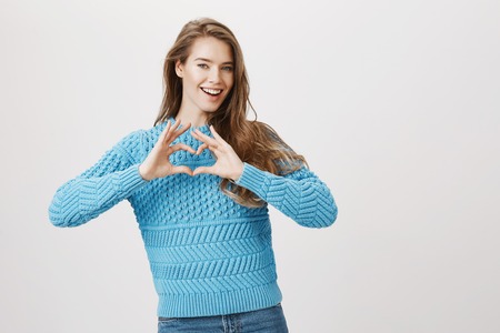 Cute and tender woman with blond hair, wearing fashionable sweater and showing heart sign near chest while smiling happily, expressing excitement and cheer, over gray background.の写真素材