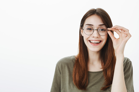 Girl ready to start working on new chapter of book. Indoor shot of creative attractive young woman in trendy round glasses, holding rim and smiling broadly, standing over gray backgroundの写真素材