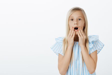Girl sees adorable doll in store, asking mother to buy it. Portrait of amazed and surprised cute fair-haired daughter, dropping jaw, saying wow and holding hands near opened mouth over gray wallの写真素材