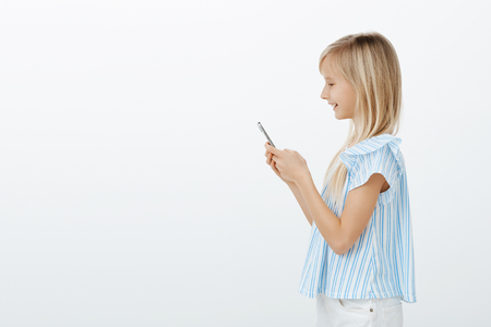 Profile portrait of positive joyful fair-haired woman in trendy blue blouse, holding smartphone and smiling at screen while writing message or watching cartoons while waiting for dinner over gray wallの写真素材