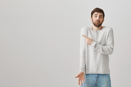 Uncertain charming caucasian guy with beard and moustache shrugging and pointing left while being...