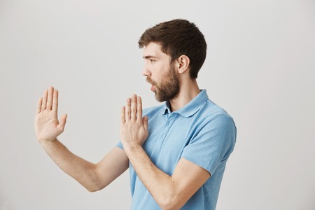 Profile portrait of playful cute bearded guy holding raised palms like in karate, standing in defense pose over gray background. Childish spouse fools around after watching film about ninjaの写真素材