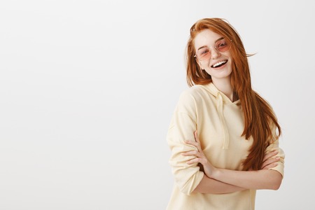 Indoor shot of creative good-looking european girl with red hair wearing trendy glasses and stylish yellow hoodie, smiling broadly, half-turned with crossed handsの写真素材