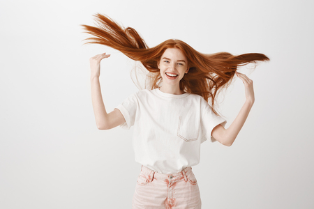 Girl raising ginger hair in air joyfully, having fun and playing with mates. Portrait of carefree happy woman in stylish t-shirt and pink jeans, playing with red strands and smiling broadly at cameraの写真素材