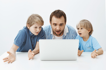 Father and sons watching video tutorial, wanting make surprise for mother. Portrait of clueless confused dad and boys sitting at table and looking at laptop screen, being unaware and questionedの写真素材