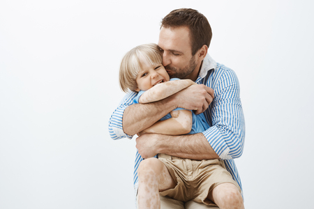 Father loves his little son like no one else. Cute good-looking caring dad hugging and kissing child in cheek, feeling happy spending time with kid, standing over gray wall, taking care of familyの写真素材