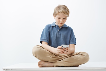 Guy feeling intense playing favorite game in smartphone. Serious pretty little boy with blond hair sitting on floor with feet crossed, holding telephone and gazing at device screen, messagingの写真素材