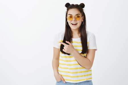 Surprised excited and happy young caucasian female in stylish clothes and sunglasses, dropping jaw and smiling broadly while pointing left with index finger and gazing at cameraの写真素材