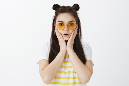 Portrait of shocked and intrigued good-looking girl in yellow sunglasses and t-shirt, holding palms on face, gasping and staring at camera while hearing incredible news or gossipの写真素材