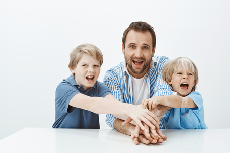 Portrait of happy playful family sharing joyful moments, expressing unity and happiness. Father and sons holding hands together while working in team over grey backgroundの写真素材