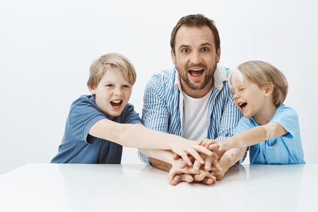 Indoor shot of joyful attractive boys and father sitting at table, holding hands, laughing and shouting from happiness. Dad teaming with sons to make present for mom, sharing positive moments togetherの写真素材