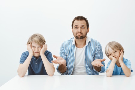 Portrait of clueless nervous father-in-law sitting with two cute boys at table, shrugging and being questioned how lift mood while guys are bored and indifferent over gray wallの写真素材