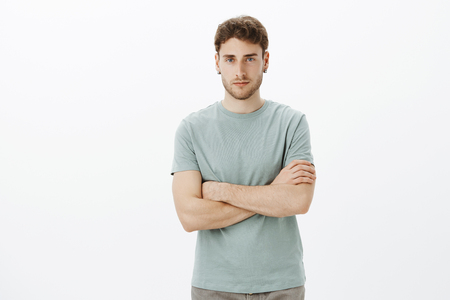 Indoor shot of confident good-looking european guy with fair hair in casual t-shirt, holding hands crossed on chest and looking with serious expression at camera, being self-assured and determinedの写真素材