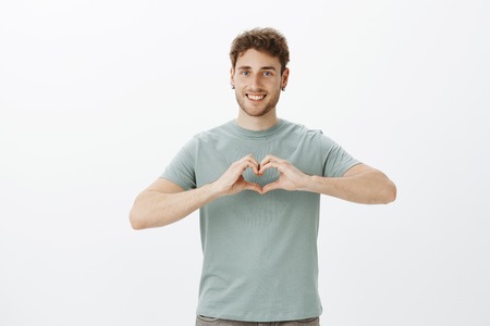 Portrait of happy friendly-looking positive guy in earrings showing heart gesture over chest and smiling broadly while expressing affection or romantic emotions over gray wallの写真素材