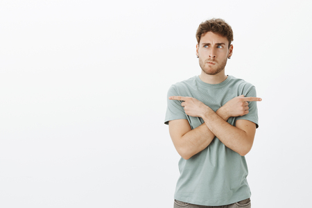 Portrait of confused european male model in earrings, smirking and frowning while thinking, crossing hands and pointing left and right while choosing direction, standing and deciding where to goの写真素材