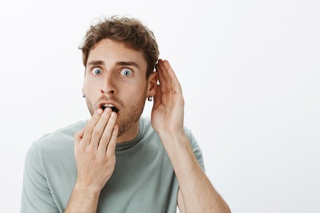 Portrait of shocked and amazed handsome guy with fair hair, dropping jaw, covering opened mouth with palm and holding hand near ear, hearing rumor or gossipの写真素材