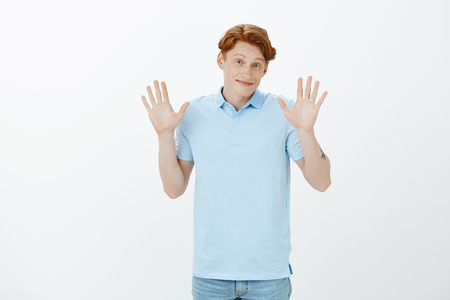 Carefree good-looking european student with red hair, raising palms in surrender and smiling broadly, giving up or trying to apologize for making fault not on purpose, standing over gray wallの写真素材