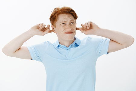 Annoyed intense guy with red hair in trendy shirt, covering ears with index fingers, grimacing and frowning, looking up, being displeased with annoying noise from upstairsの写真素材