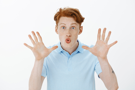 Guy telling horror story by fire during vacation with friends. Cute good-looking redhead male with freckles, folding lips showing palms while wanting to scare someone, standing intense over grey wallの写真素材