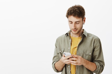 Handsome carefree curly-haired man with beard, holding smartphone and gazing at screen of device while listening music in earphones, enjoying good sound over grey wallの写真素材