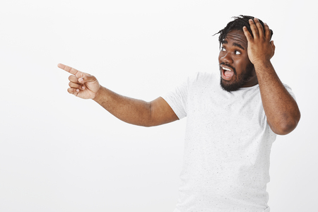 Portrait of shocked impressed dark-skinned guy in white t-shirt, stretching hand, looking and pointing left while holding palm on hear, being stunned with accident, showing anxiety over grey wallの写真素材