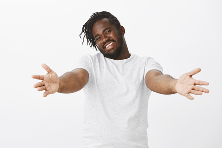 Friendly loving plump african man with beard, stretching hands towards camera to cuddle, smiling broadly and expressing happy and caring attitude, standing over white wallの写真素材