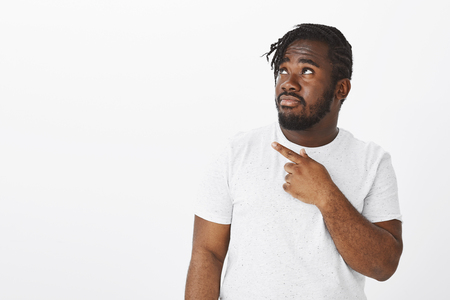 Indoor shot of intense worried dark-skinned male model with beard and moustache, looking and pointing at upper left corner, being scared or displeased with huge spider on ceiling over white backgroundの写真素材