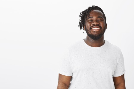 Waist-up shot of happy positive dark-skinned guy in casual t-shirt, smiling broadly and friendly at camera while posing for advertisement over white background, feeling carefree and joyfulの写真素材