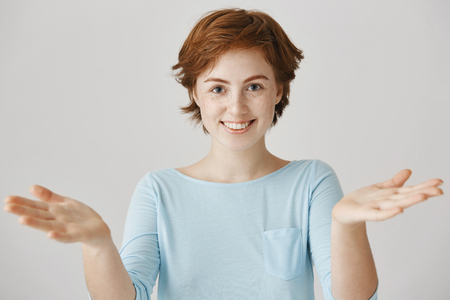 Portrait of attractive redhead caucasian girlfriend with cute freckles pulling palms towards camera as if weighing something or wanting to hug friend, standing over gray wall with broad smileの写真素材