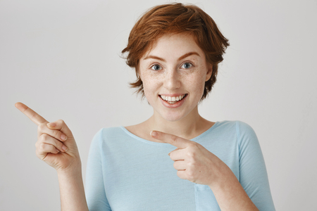 Loot at that direction. Beautiful caucasian redhead with freckles pointing left with forefingers and smiling broadly, being up to something funny and good, standing over gray background.の写真素材