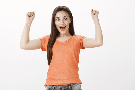 Sudden success sweep off fit. Studio shot of happy triumphing cheerful woman in orange t-shirt, raising clenched fists and yelling yes from happiness, being winner and champion over gray backgroundの写真素材