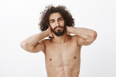 Waist-up shot of confident beautiful hispanic bearded guy with curly hair, holding hands behind neck, standing naked with tattoo on chest, smiling with satisfaction, being carefree and flirtyの写真素材
