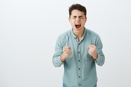 Cannot handle feelings inside. Portrait of desperate intense angry european guy, screaming and clenching fists, standing with closed eyes and bending towards camera, being pissed off and fed upの写真素材