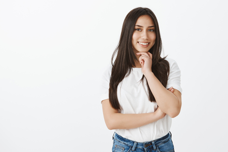 Portrait of creative feminine girl with beautiful long hair in jeans and white t-shirt, holding hand on chin and smiling at camera with curiosity and interest having great plan or ideaの写真素材