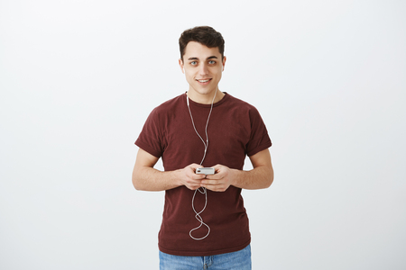 Which song listen during walk to work. Portrait of happy handsome boyfriend in casual red t-shirt, holding smartphone and earphones, looking satisfied and joyful at camera while hearing new albumの写真素材