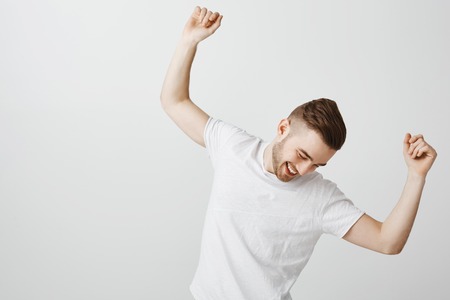 Happy and pleased good-looking guy with beard in white t-shirt celebrating with joy accomplished work dancing and sliding of floor with raised hands looking down smiling broadly with satisfactionの写真素材