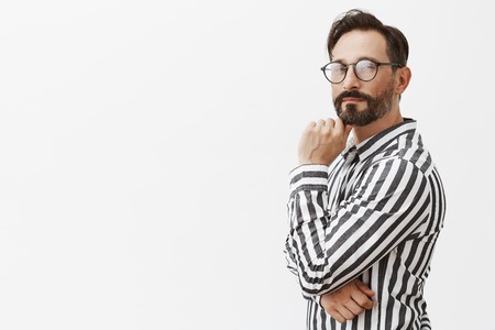 Hmm, nice idea, I will use it. Thoughtful strategist in glasses and striped shirt, standing in profile and smirking at camera, rubbing beard while thinking, having great plan over gray backgroundの写真素材