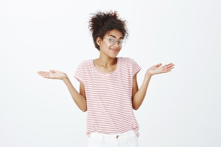Indoor shot of indifferent happy young woman in glasses and striped t-shirt, shrugging with spread...