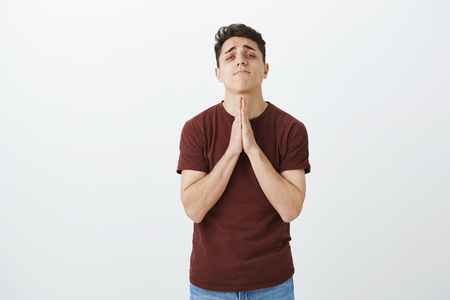 Please, help me I beg you. Cute nervous attractive guy in casual outfit, holding hands in pray and staring with lifted chin at camera while apologizing or wanting favour, standing over gray wallの写真素材
