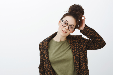 Waist-up shot of attractive modern caucasian girl in black stylish glasses and leopart coat over casual t-shirt, touching hairbun, tilting head, posing over gray background, relaxed and confidentの写真素材