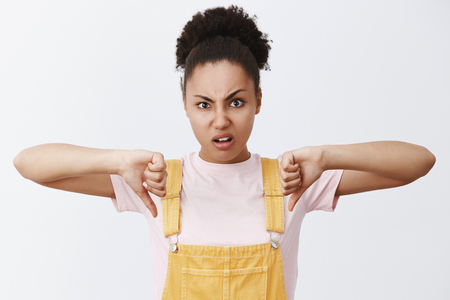 Nah, it is dislike. Portrait of displeased and disappointed cute African American girlfriend in trendy yellow overalls, showing thumbs down and frowning from disgust after seeing awful movie in cinemaの写真素材
