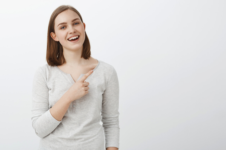 Go see yourself. Portrait of happy opimistic good-looking young woman with short brown hair and cute ear smiling joyfully with opened mouth pointing at upper right corner suggesting vising nice placeの写真素材