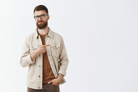 Indifferent displeased adult european male entrepreneur in beige jacket over brown t-shirt and glasses with sick long hipset beard pointing right frowning from scorn and disdain, unimpressed dislikingの写真素材
